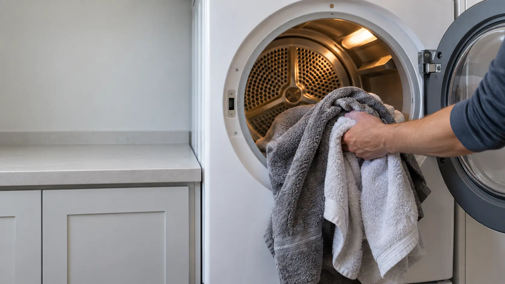 A hand holding damp towels just taken from an open front-load dryer in a US home laundry room, the drum interior visible.