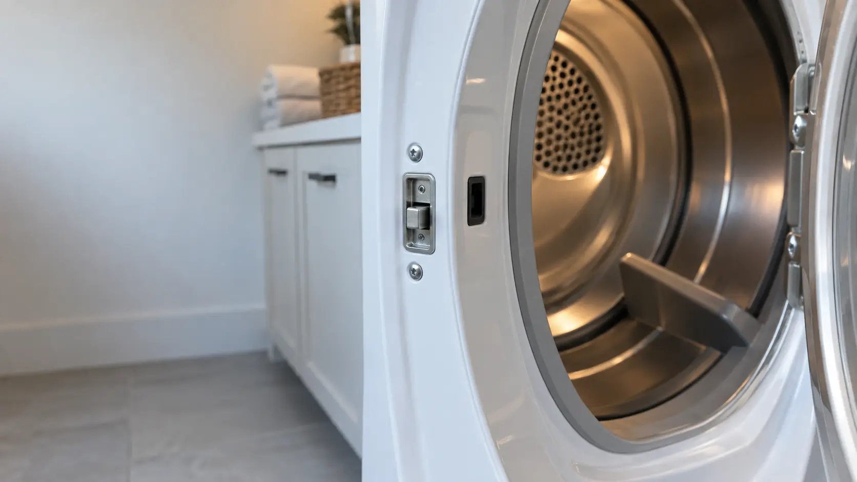 The open door of a front-loading dryer seen close at the latch side, the strike and the small door switch opening on the cabinet front.