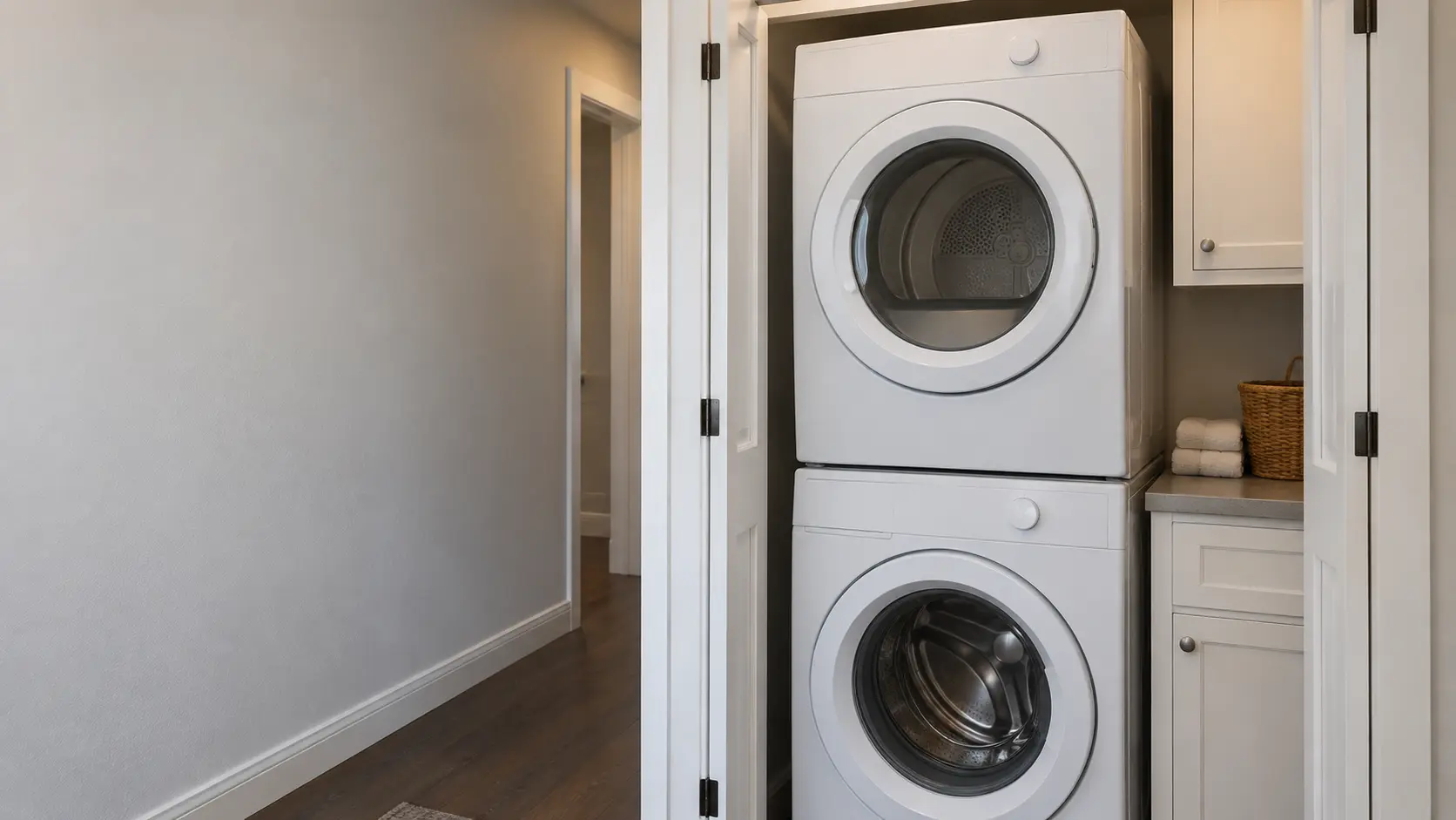 A white front-load washer with a matching dryer stacked on top, fitted into a narrow laundry closet off a US hallway with bifold doors folded open.